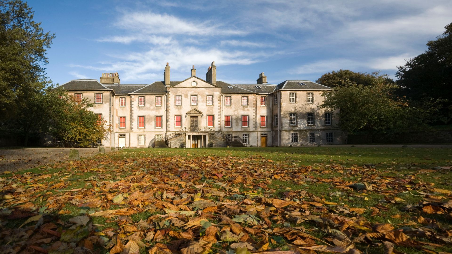 The front of a symmetrical Palladian house with lots of windows and a large grassy area covered in fallen autumn leaves leading up to the front door, perfect for Halloween events.