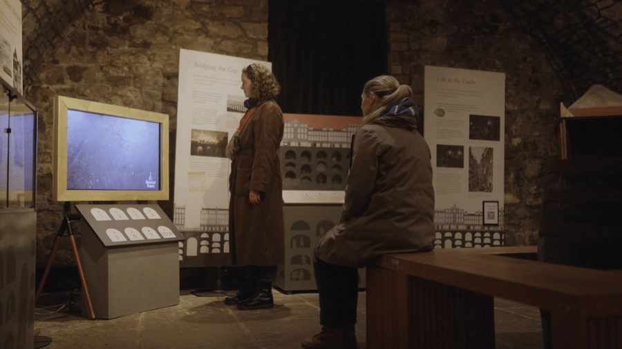Two people, one standing and one sitting, watch The Vaults Revealed: a TV with an interactive interface in the Edinburgh Vaults Museum.