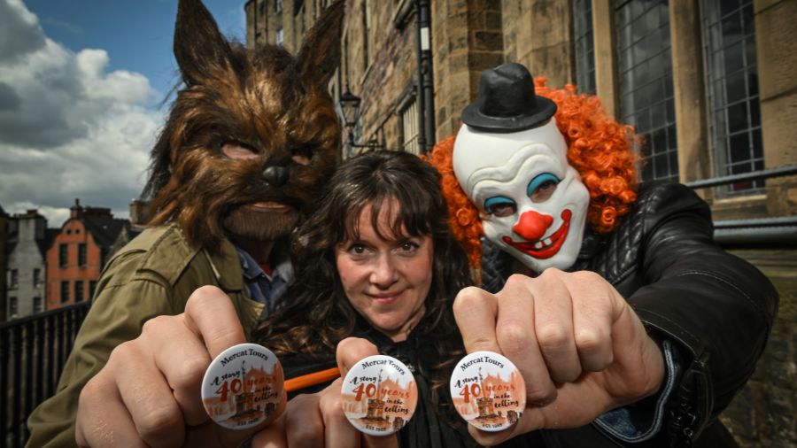 Three costumed figures, a wolf, a cloaked Mercat Storyteller, and a clown, pose playfully with celebratory fortieth birthday button badges outside the stone buildings on Victoria Terrace in Edinburgh