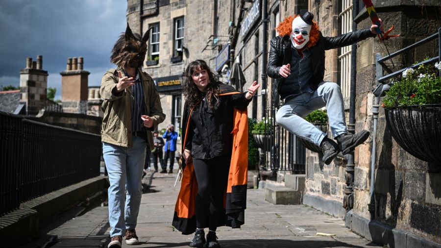 Three people in costume walk down the historic Edinburgh Victoria Terrace, with a wolf on the left, a cloaked Mercat Tours storyteller in the centre, and a clown leaping playfully in the air on the right.