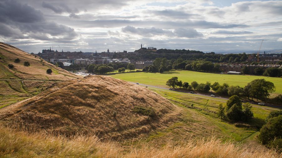 A photo taken from a hill in Holyrood Park, looking out over lots of green space and towards the city centre. Edinburgh Castle is visible on a hill not too far away.