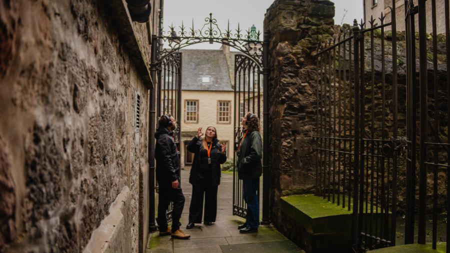 A Mercat Storyteller gestures up at the iron gated archway leading into Tweeddale Court as 2 guests look on.
