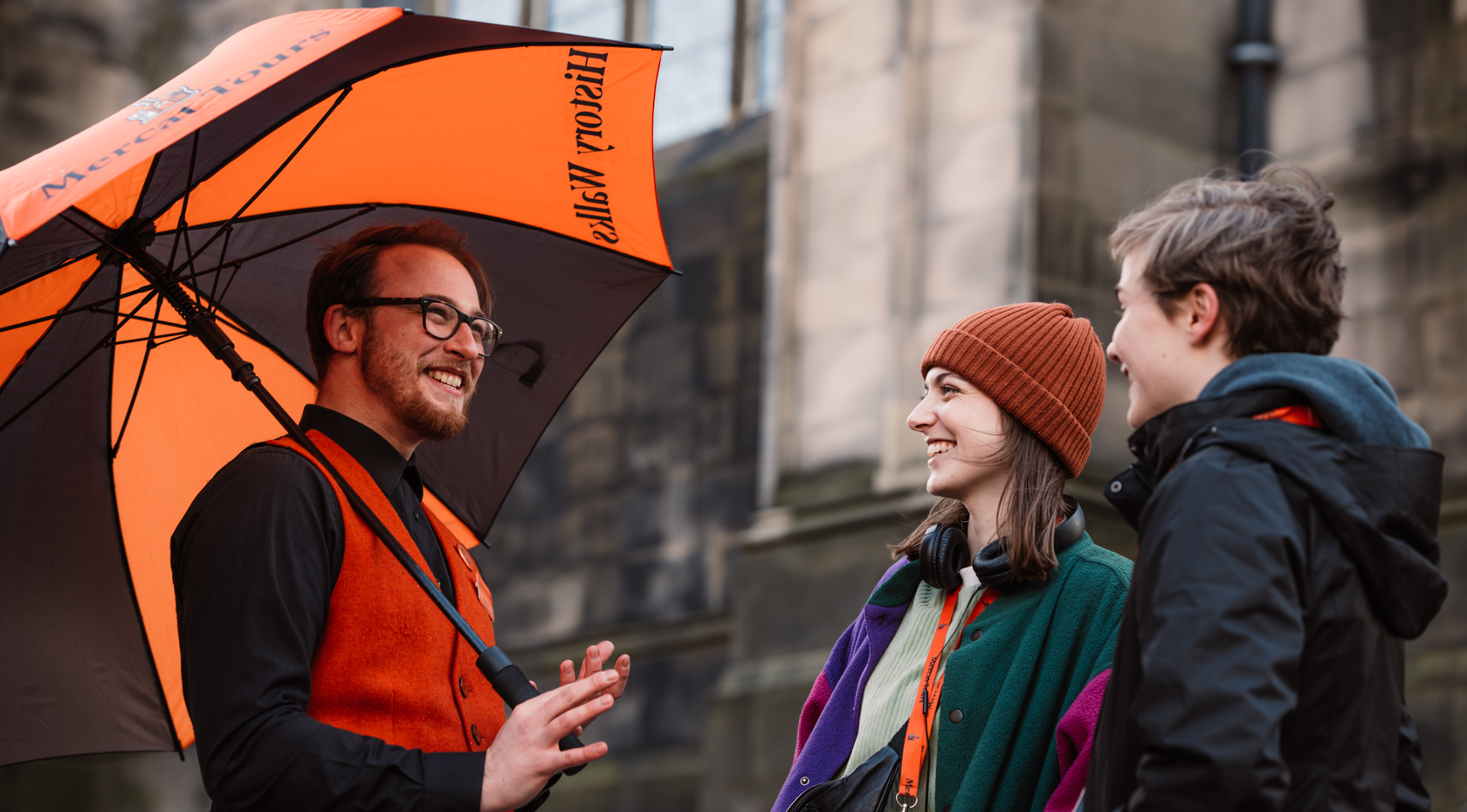 A Mercat Tours Support Team member holding a brand umbrella, talking to two visitors wearing TourTalks and headphones.