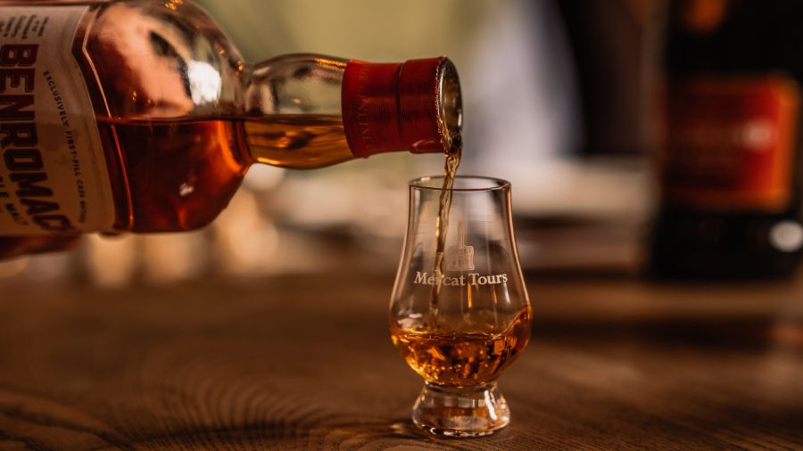 A hand pouring whisky into a small Glencairn glass etched with the Mercat Tours logo.