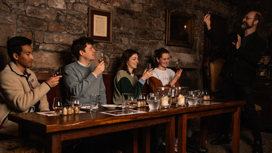 Four people sit at a wooden table in the candlelit Megget’s Cellar, being led through an Edinburgh whisky tasting by a Mercat Storyteller.