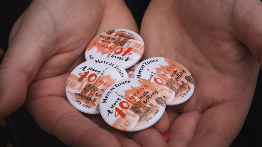Hands holding four buttons with an illustration of the Mercat Cross and text that reads 'Mercat Tours; A story 40 years in the telling; est. 1985.