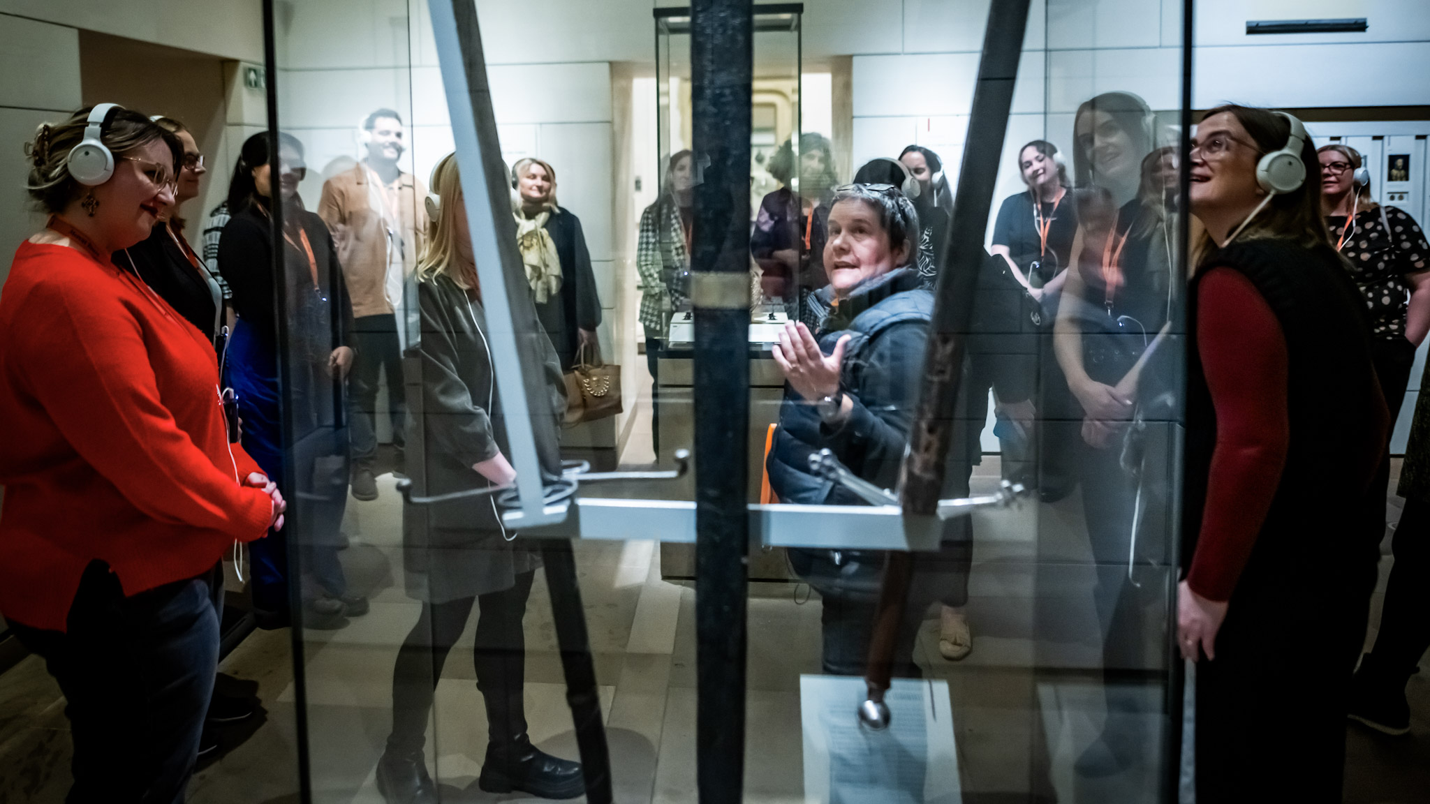 A Mercat Storyteller seen through the display case of a large ceremonial sword, surrounded by visitors wearing white headphones. 