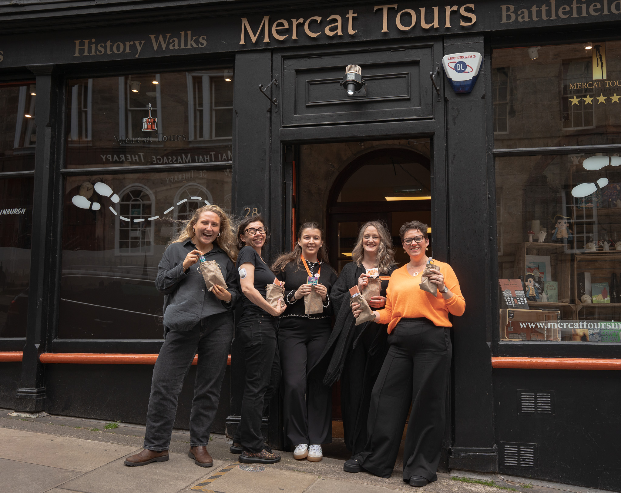 5 women stand in front of a black building with the name Mercat Tours above the door. They smile while holding burlap bags with various sticking out, like chocolate bars. 