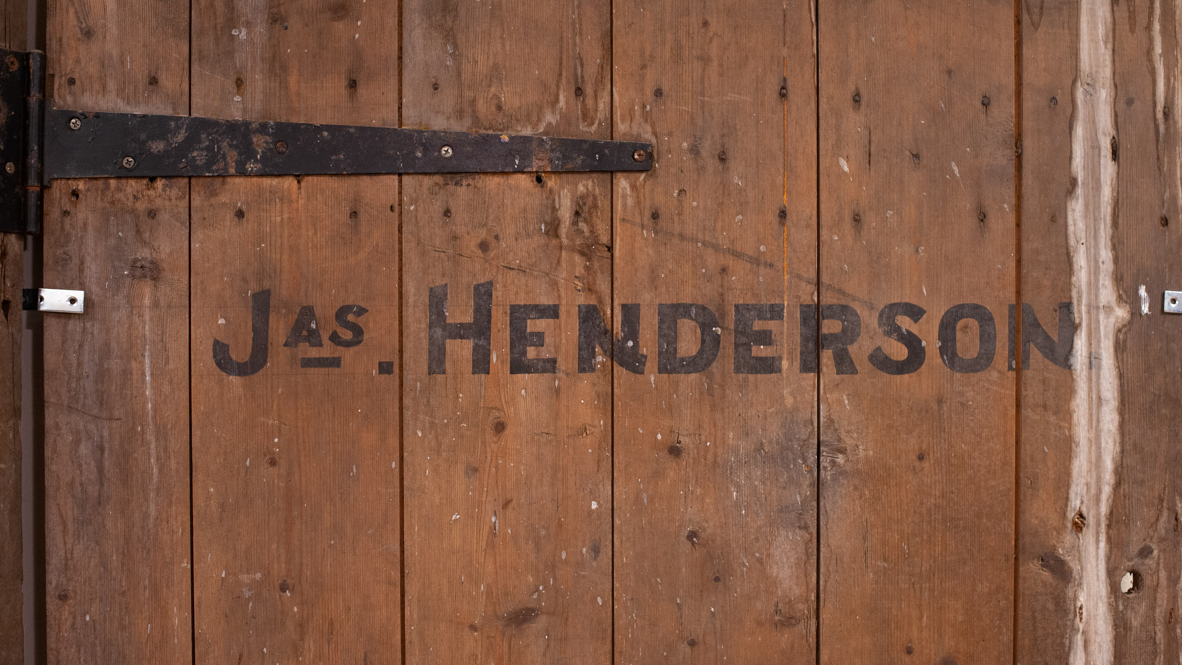A close-up of a wooden door with old black hinges, water damage, and large black letters that read ‘Jas. Henderson.’