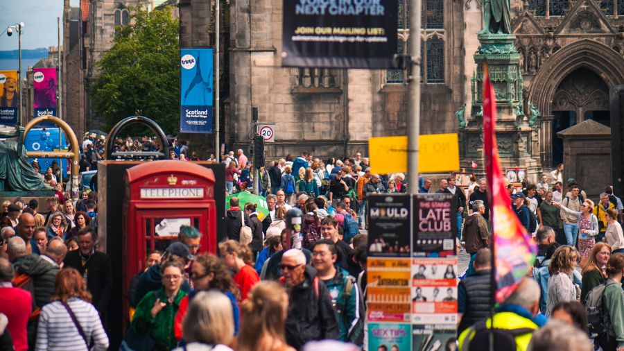 The Royal Mile in Edinburgh on a sunny day, full of huge crowds of people and posters for Fringe Festival shows.