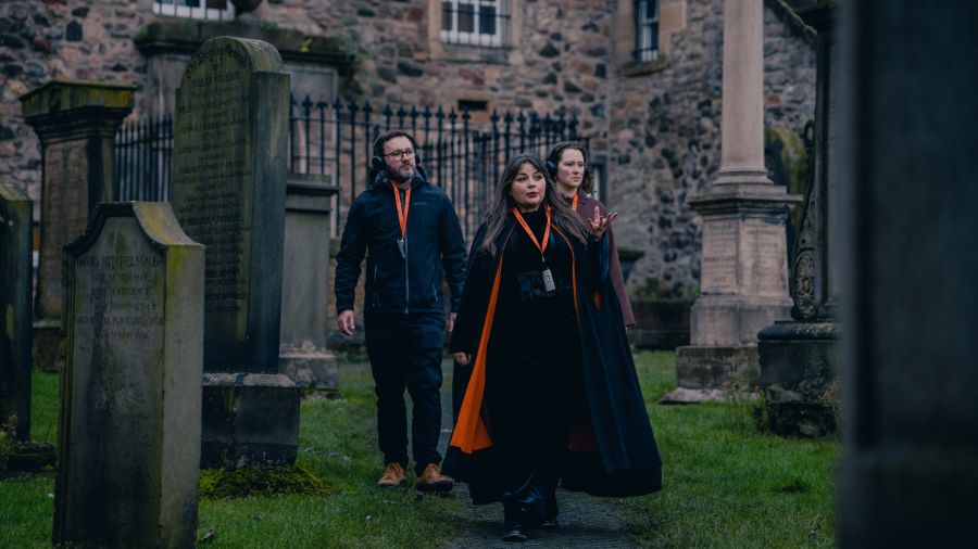 A Mercat Storyteller in a black cloak leads 2 visitors down a path in the Canongate Graveyard on a ghost tour of Edinburgh.