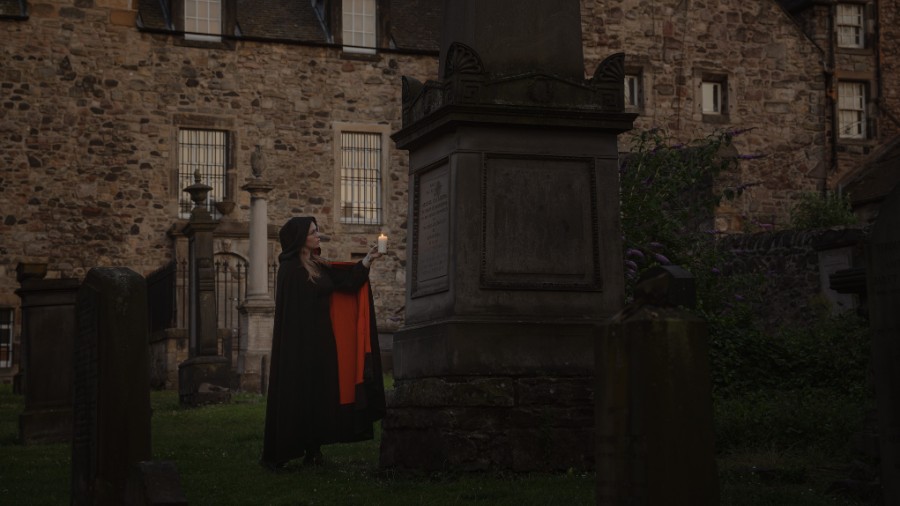 A Mercat Stroyteller in a black cloak with orange linings stands in Canongate Kirkyard, raising a candle to a tall grave during an Edinburgh Halloween graveyard tour.