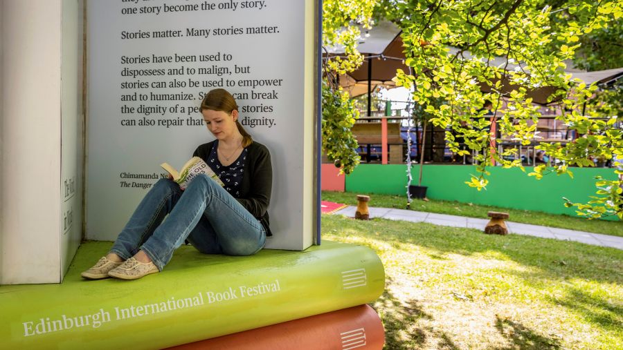 A woman sits on a book display while reading, the bench labelled with ‘Edinburgh International Book Festival’.