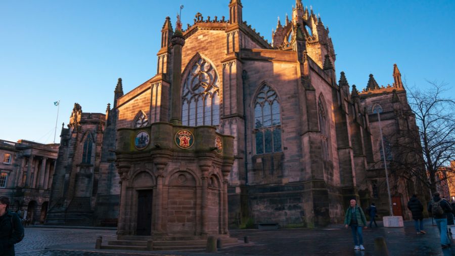The Edinburgh Mercat Cross on the Royal Mile with St Giles’ Cathedral in the background against a bright blue sky.
