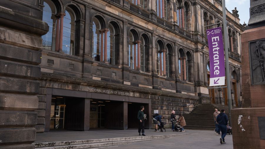 The front of the National Museum of Scotland in Edinburgh, a Victorian building with a purple Entrance banner on a flagpole.