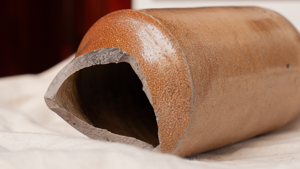 A close-up of the broken neck of a stoneware bottle resting on canvas fabric