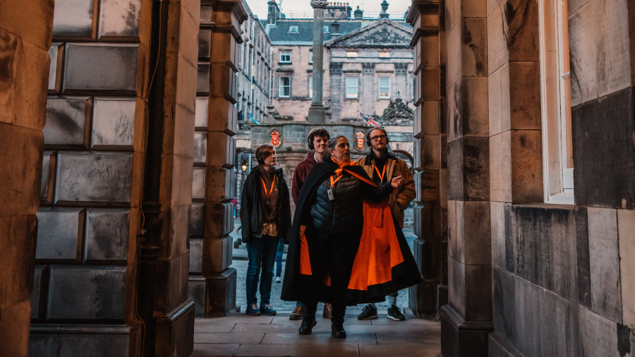 A Mercat Storyteller leads a group of 3 people on a walking ghost tour of Edinburgh, with the Mercat Cross behind them, framed by an archway.