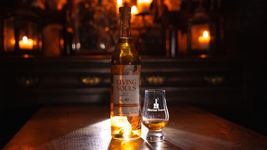 A backlit bottle of 40-year-old Living Souls whisky next to a Glencairn glass etched with the Mercat Tours logo, in front of a sideboard moodily decorated with lit candles.