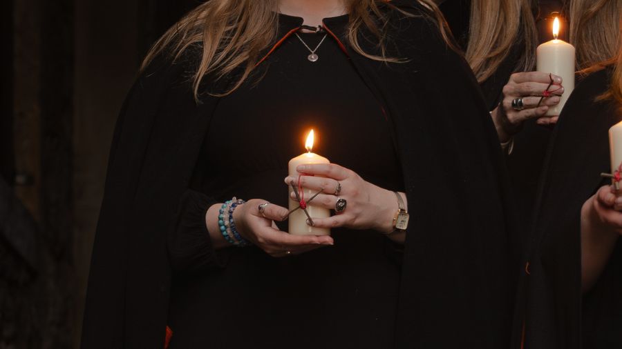 The hands of a Mercat Storyteller holding a small rowan cross tied together with red cord and a lit candle.