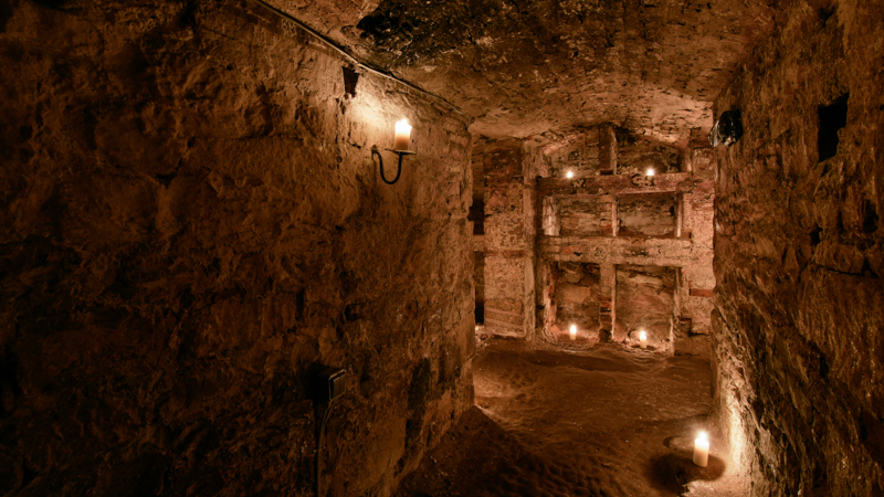 A candlelit corridor of the Blair Street Underground Vaults, ending in a wall of stone storage spaces.