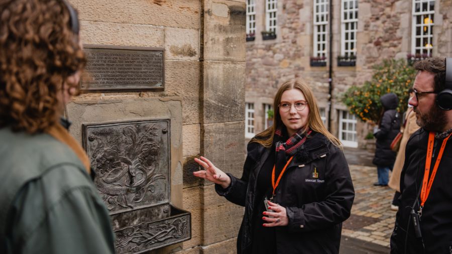 A Mercat Storyteller wearing a Witches of Scotland tartan scarf gestures to a fountain head in a wall as two visitors look on.