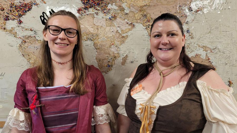Two women in period dress pose in front of a map of the world, one with stiches around her throat and the other with a loop of rope around her neck.