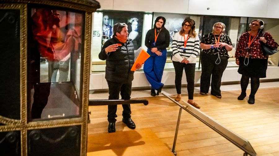 A Mercat Storyteller speaks to a group of visitors at the National Museum of Scotland, gathered around a large, covered chair with poles on either side. 
