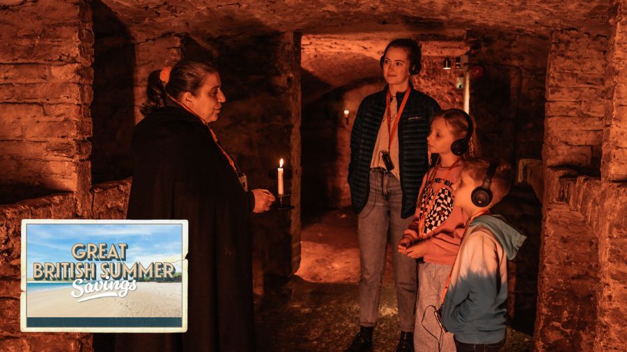 A Mercat Storyteller leading an adult and two kids on a kids ghost tour in Edinburgh, inside the Edinburgh vaults. The logo, in the form of a postcard, for the Great British Summer Savings scheme is in the lower left-hand corner.
