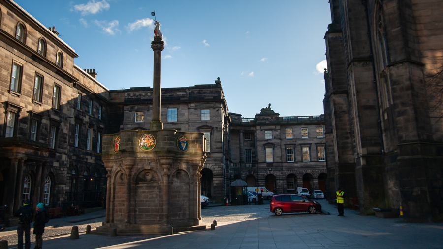 The Edinburgh Mercat Cross, a monument with a column topped by a unicorn holding a shield and a Scottish flag, surrounded by Georgian buildings and St Giles Cathedral. 