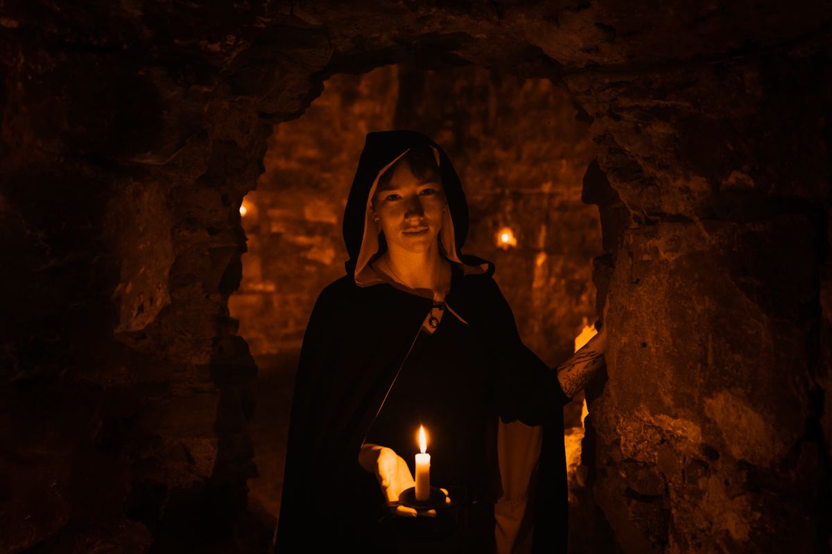 A Mercat Tours Storyteller holding a lit candle, walking through a stone archway in the Blair Street Underground Vaults, Edinburgh.