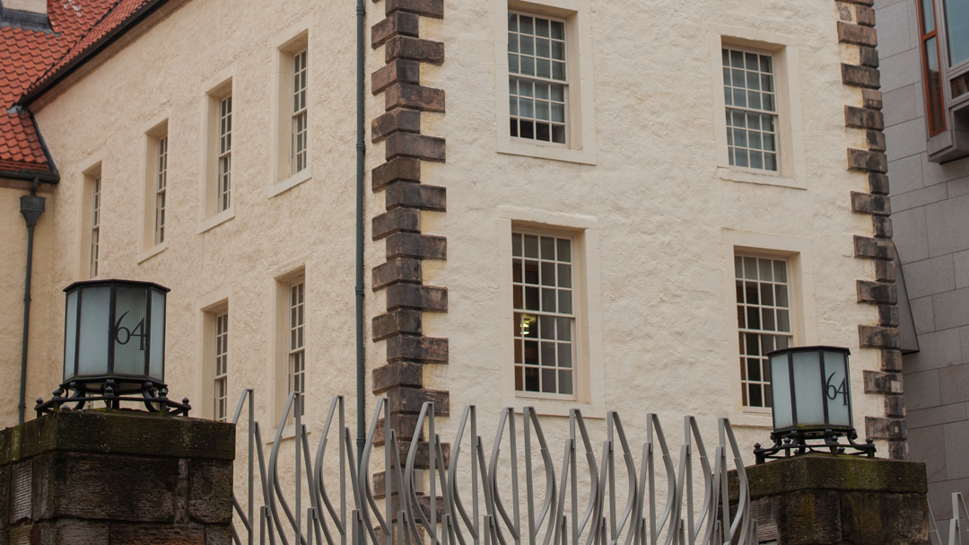 A yellow stucco house with stone details at its corners, lots of paned windows, an orange roof, and a modern metal gate flanked by two stone pillars topped by lanterns reading the number 64.