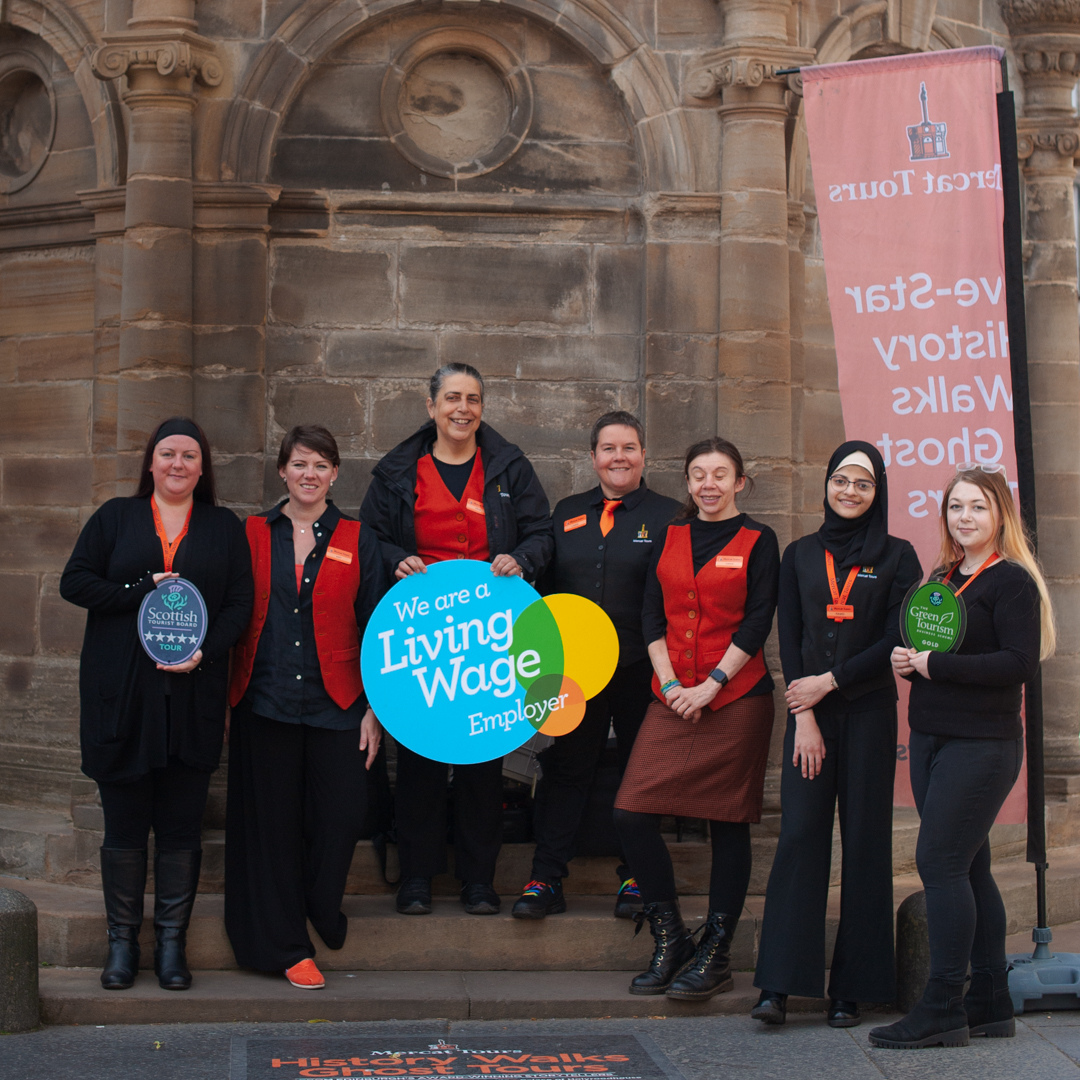 Seven Mercat Tours team members standing in front of the Mercat Cross, with a banner behind. Three individuals are holding the 5-star Scottish Visitor Attraction Award, the Living Wage Employer logo and the Green Tourism GOLD Award.