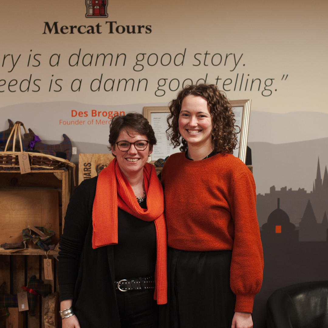 Two women, both dressed in orange and black, smile directly at the camera in a reception area. They are stood in front of a Mercat Tours logo and partial quote - 'History is a damn good story. What it needs is a damn good telling.'