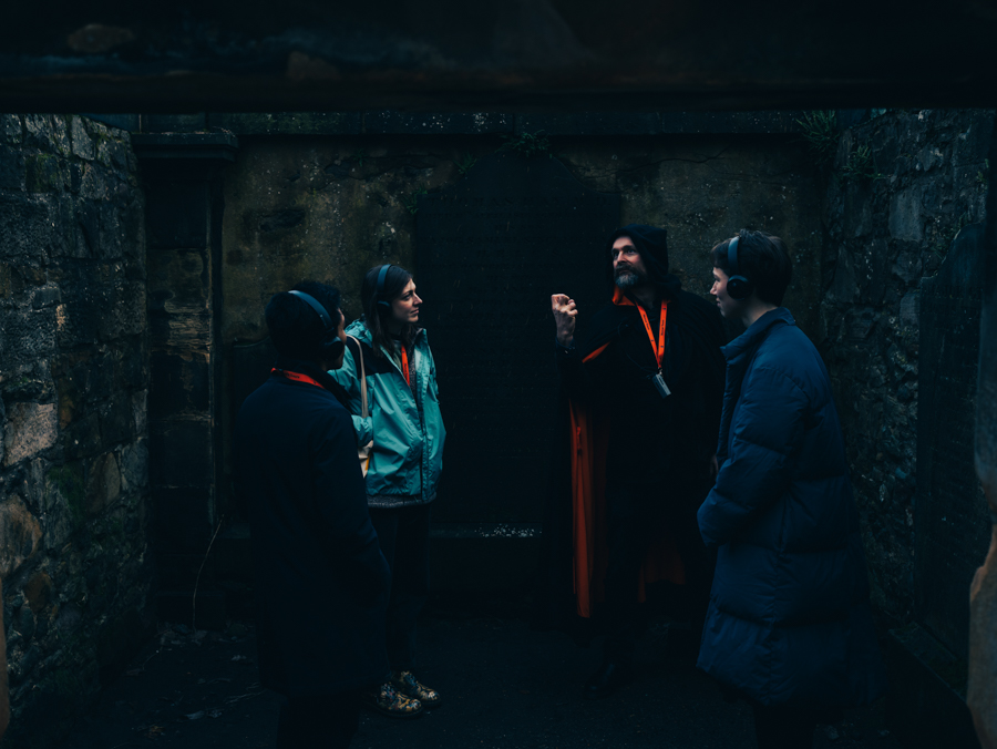 A Mercat Storyteller in a black cloak stands inside a mausoleum in Canongate Kirkyard with a group of engaged visitors.