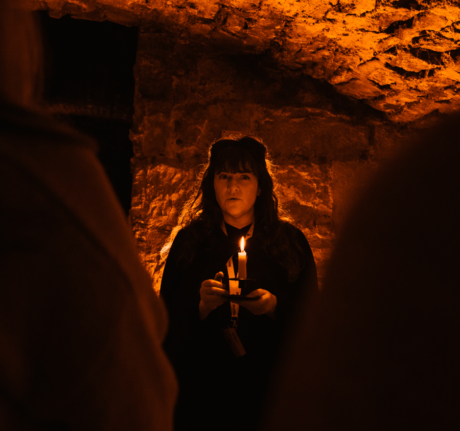 A Mercat Storyteller in a black cloak stands in a corner of the Blair Street Underground Vaults, holding a candle and framed by visitors.