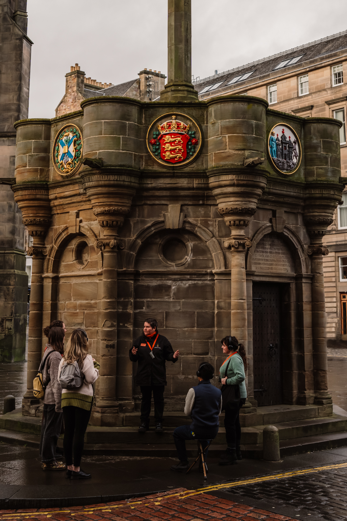 A Mercat Storyteller in a black jacket stands at the Mercat Cross with a group of engaged visitors.