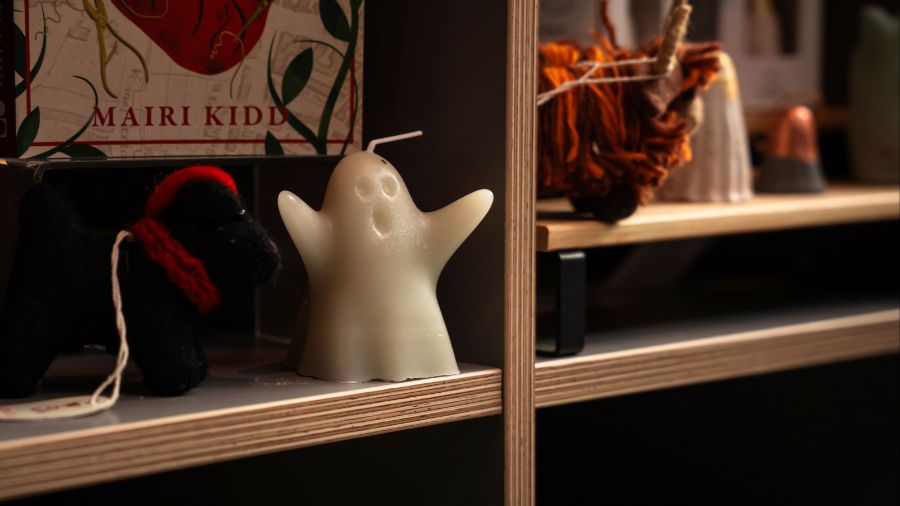 A white candle in the shape of a ghost sits on a shelf next to a wool terrier ornament and other souvenirs.