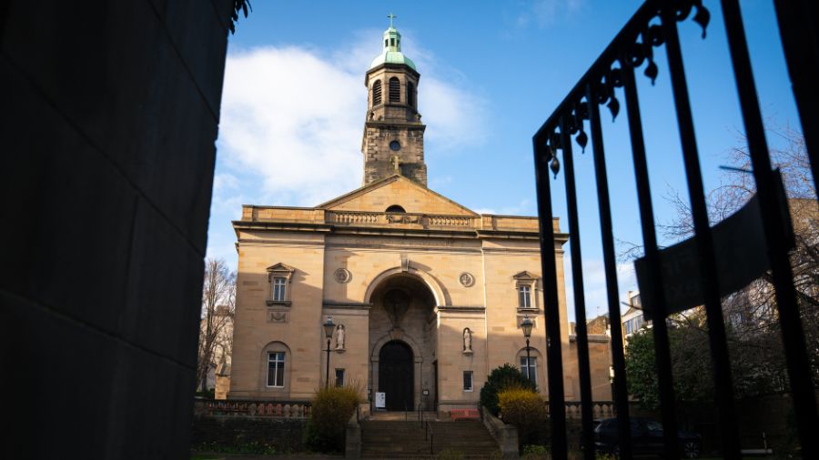 The front of the St Patrick's church sunlit large light stone with tower behind green dome and cross at top, with blue sky behind.