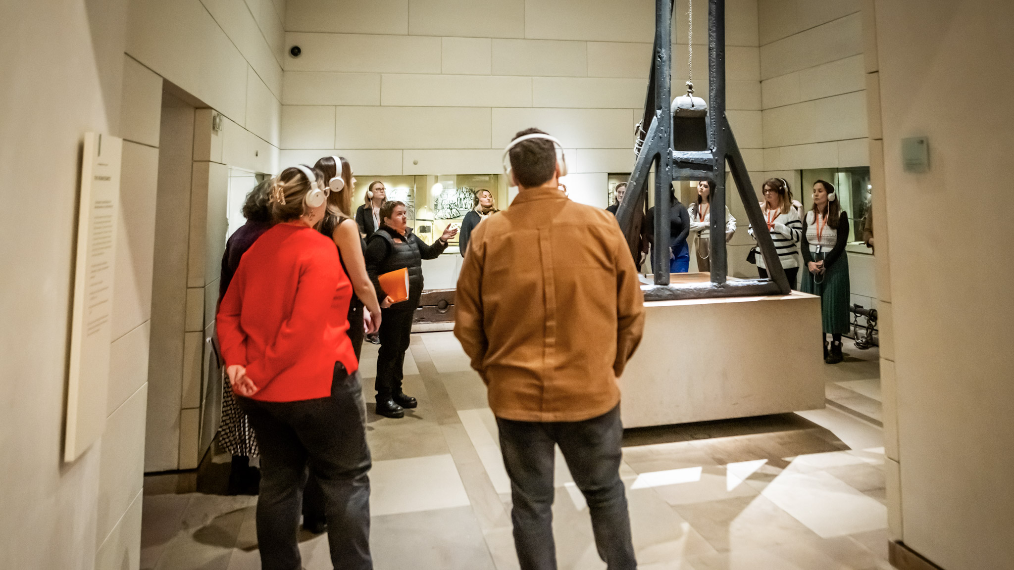A Mercat Storyteller standing next to a wooden guillotine at the National Museum of Scotland, surrounded by visitors wearing white headphones. 