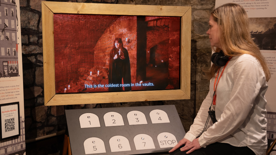 A woman sits next to an audio-visual display featuring eight buttons and a TV, which displays a Mercat Storyteller holding a candle in the Blair Street Underground Vaults and the subtitles ‘This is the coldest room in the vaults.’