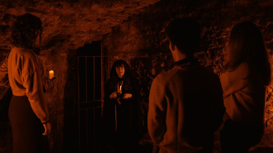 A Mercat Storyteller in a hooded black cloak shares stories with three guests in a room of the Edinburgh underground vaults. 