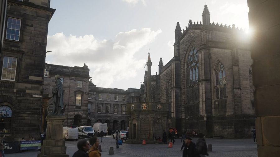 The Mercat Cross seen at a distance with St Giles Cathedral behind it, a Mercat Tours team member with an orange and black umbrella standing in front of it.