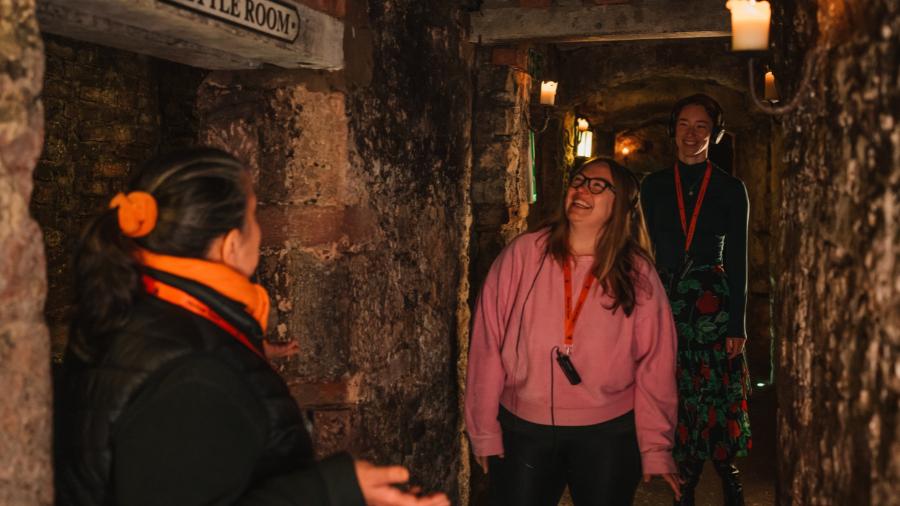 A Mercat Storyteller leading a pair of smiling and laughing women down a corridor of the Edinburgh underground vaults, on a history tour of Edinburgh.